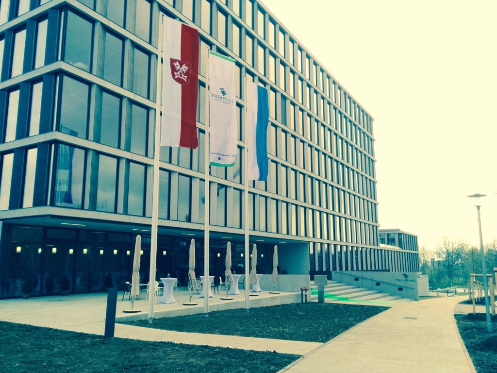 Foto vom Eingang zur TechBase Regensburg, vorne drei Fahnen und ein grüner Teppich zur Feier der Eröffnung des Gebäudes